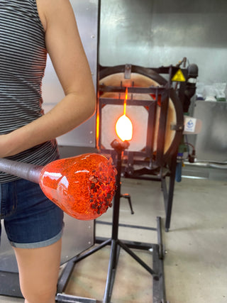 Close up image of glass blower holding hot molten glass at the end of a pole in the hot shop.