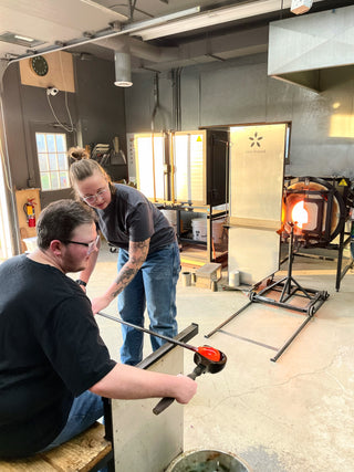 Two students working together to create a glass piece in a glass blowing class