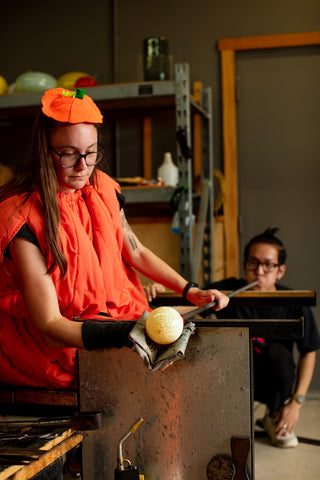 Glass artist in a pumpkin costume using tools to shape molten glass