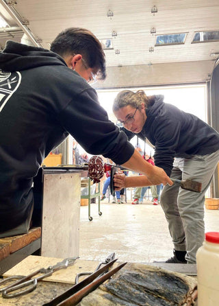 Two artists demonstrating how to work in the hot glass studio in front of a crowd